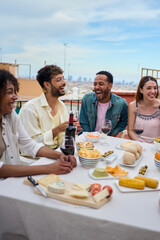 Vertical. Laughing group multiracial young friends enjoying lunch together outdoors. Cheerful people gathered celebrating snack party on rooftop. Millennial generation of friendship on vacation. 