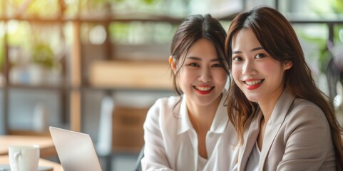 Wall Mural - two young Asian women smiling and sitting in front of their laptops, with a modern office space as the background.