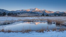 A Tranquil Winter Scene Featuring A Partially Frozen Pond, Golden Sunlit Snow-capped Mountains, And A Clear Blue Sky At Dawn.