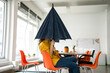 © Westend61 - Creative businesswoman holding folded umbrella covering face at workplace