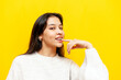 young pensive asian girl with braces flirts and thinks sexually over yellow isolated background, excited korean girl dreams and imagines and points finger at lip