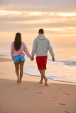 Fototapeta Sport - Rear View Of Couple In Casual Clothing On Vacation Holding Hands Walking Along Beach Shore At Dawn