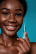 Smiling African American girl with a syringe in her hand on a blue background. Skin care, beauty injections, anti age. Vertical orientation.