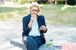 © okrasiuk - Middle-aged business woman eating sandwich burger with salad and using phone sitting on bench in the park while having lunch break. Remote work,business, freelance,blogging,social media concept