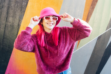 Wall Mural - Hipster young woman with pink hair and sunglasses in magenta fluffy sweatshirt and bucket hat posing on the wall background. Urban street fashion. Mono color look. Gen Z, millennials self expression.