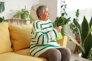 Wall Mural - Senior gardener with plant and spray water bottle sitting on sofa at home