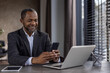 © Liubomir - Confident African American businessman in a suit smiling while using his smartphone and laptop in a modern office setting.