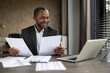 © Liubomir - African American businessman in a suit smiles while reviewing papers in an office setting with a laptop and other documents on his desk.