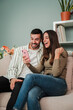 © Jose Calsina - Vertical. Couple having fun using a mobile phone to browse on internet at living room sofa. Two friends betting online with a cellphone app. Boyfriend and girlfriend searching or booking an apartment