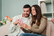 © Jose Calsina - Caucasian couple having fun using a mobile phone to browse on internet at living room sofa. Two friends betting online with a cellphone app. Boyfriend and girlfriend searching or booking an apartment