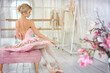 Adorable young blond female ballet dancer with bare back in pink tutu siting on bench and putting shoes on during training in white class room. Elegant barefoot, ballerina. Dance, art concept.