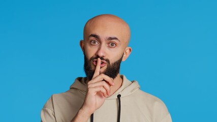 Wall Mural - Middle eastern man doing hush silence symbol in studio, asking to keep a secret over blue background. Arab person creating mute gesture to be quiet in front of camera. Camera 2.