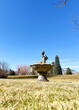 Large  cement birdbath fountain with pedestal sits in a grassy yard with mountain & tree scenery beyond. A statue figure of a girl washing clothes sits atop the fountain. Blue sky is above. Text space