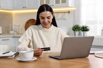 Canvas Print - Happy young woman with credit card using laptop for shopping online at wooden table in kitchen