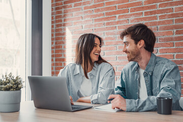 Happy millennial couple sit at table at home browsing web on laptop shopping online together, smiling young husband and wife work on computer at desk pay household bills or taxes in internet banking.