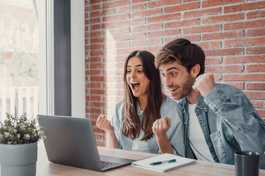 Overjoyed millennial couple sit at desk look at laptop screen feel euphoric with online win, excited happy young husband and wife read good news, get unexpected notification on computer, luck concept.