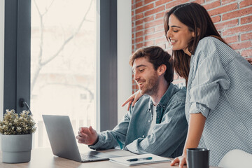 Happy young family couple bonding laughing using laptop looking at screen sit at table, smiling husband and wife buying choosing goods online doing internet shopping order with delivery at home.