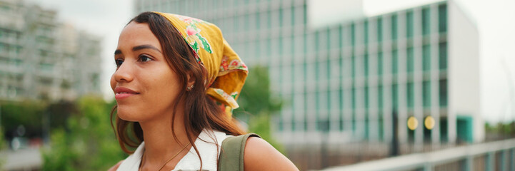 Wall Mural - Сlose up of cute tanned woman with long brown hair wearing white top and yellow bandana walks on bridge with backpack on her shoulder and looks at modern city, Panorama