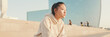 © Andrii Nekrasov - Asian girl in sportswear resting after workout sitting on steps on modern buildings background at sunrise, Panorama