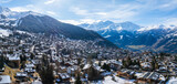 Fototapeta  - Aerial shot of Verbier, a Swiss Alps ski town, displays chalets in a snowy setting. It shifts through seasons under a clear sky, with grand mountains behind. Perfect for winter sports fans.