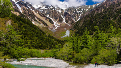Wall Mural - Two hikers beside a clear river leading towards high, snowy mountain peaks behind