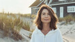 an attractive woman in her early fifties wearing a white hoodie. She has shoulder length brown hair and is standing on the beach next to a house. She looks confident with a gentle smile on her face