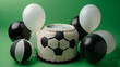 A sports-themed birthday cake resembling a soccer ball, flanked by black and white balloons on a solid green background.