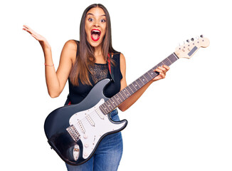 Wall Mural - Young hispanic woman playing electric guitar celebrating victory with happy smile and winner expression with raised hands