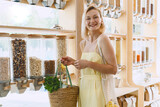 Fototapeta Tęcza - Smiling woman with basket of organic products in sustainable plastic free store. Dispensers for cereals, grains, nuts at zero waste shop.