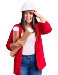 Poster - Young beautiful brunette woman wearing hardhat holding architect blueprint stressed and frustrated with hand on head, surprised and angry face