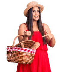 Canvas Print - Young beautiful brunette woman wearing summer hat holding picnic basket smiling happy pointing with hand and finger