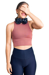 Wall Mural - Beautiful caucasian young woman wearing gym clothes and using headphones smiling and laughing with hand on face covering eyes for surprise. blind concept.