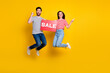 Full length photo of two crazy people jump hold sale placard raise fist direct finger empty space isolated on yellow color background