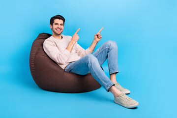 Fototapete - Full size photo of funny man wear sweatshirt denim pants sit on bean bag directing at sale empty space isolated on blue color background