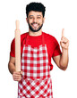 © Krakenimages.com - Young arab man with beard wearing professional baker apron holding kneading roll smiling with an idea or question pointing finger with happy face, number one