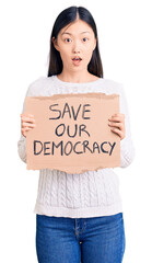 Wall Mural - Young beautiful chinese woman holding save our democracy cardboard banner scared and amazed with open mouth for surprise, disbelief face