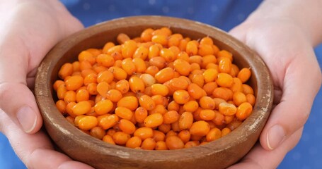 Wall Mural - Woman hands holding wooden bowl full of frozen ripe sea buckthorn berries and moving it forward. Close-up.