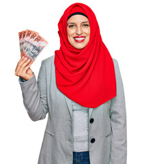 Poster - Beautiful hispanic woman wearing islamic hijab holding australian dollars banknotes looking positive and happy standing and smiling with a confident smile showing teeth