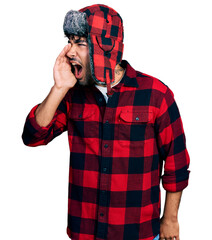 Canvas Print - Hispanic young man with beard wearing fluffy earmuff hat shouting and screaming loud to side with hand on mouth. communication concept.