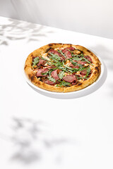 Wall Mural - Italian pizza with roast beef, arugula and spicy sauce on light background with shadows. Meat pizza with sliced roast beef on white table. Italian food in trendy menu restaurant