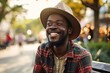 Cheerful african american man in hat and checkered shirt smiling and looking away while sitting on bench outdoors