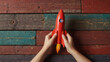 A hand holding a red crayon next to a red and yellow paper rocket