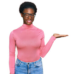 Wall Mural - Young african american girl wearing casual clothes and glasses smiling cheerful presenting and pointing with palm of hand looking at the camera.