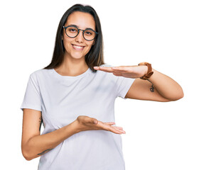 Wall Mural - Young hispanic woman wearing casual white t shirt gesturing with hands showing big and large size sign, measure symbol. smiling looking at the camera. measuring concept.