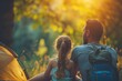 Man and girl seated by tent, bask in sunset's glow, forest around them alight in golden hour’s embrace. Parent and young one enjoy tranquil moment, back to orange tent, day wanes amidst verdant trees.