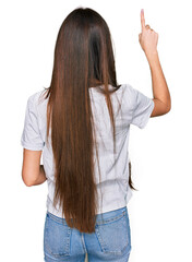 Wall Mural - Young hispanic girl wearing casual white t shirt posing backwards pointing ahead with finger hand