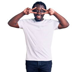 Poster - Young african american man with braids wearing casual white tshirt doing peace symbol with fingers over face, smiling cheerful showing victory