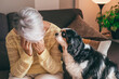 Sad senior woman sitting on sofa at home with hands on face, her cavalier king charles dog looks at her worried. Retired elderly lady and pet therapy concept