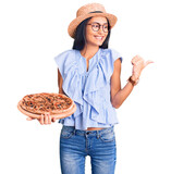 Fototapeta Koty - Young beautiful latin girl wearing summer hat and glasses holding pizza pointing thumb up to the side smiling happy with open mouth