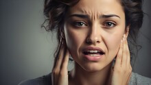 A woman with curly hair and a grey shirt is holding her jaw in pain. She has her hands on her face and her teeth are showing. Her expression is one of discomfort and distress.